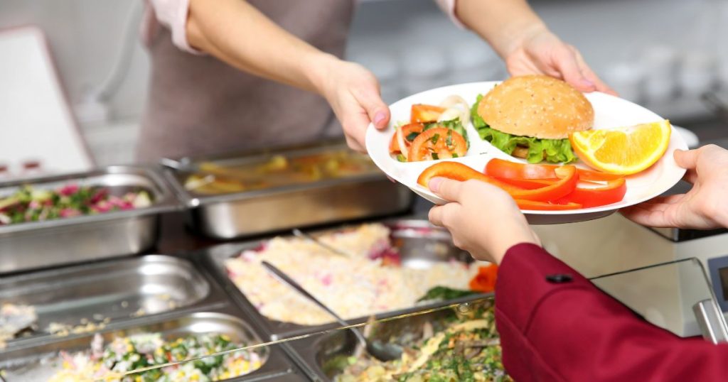 Een werknemer neemt een bord met een hamburger, rauwkost en appelsienschijfje aan van een cafetariawerknemer. 