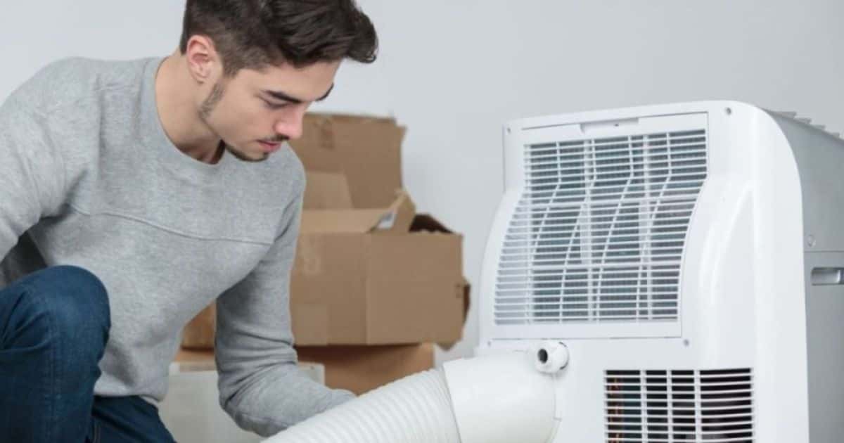 Een man installeert een mobiele airco. Op de achtergrond kartonnen dozen en een witte muur.