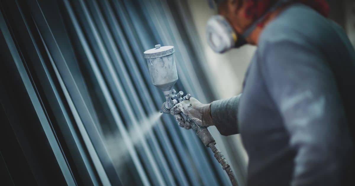 Een man in grijze kledij gebruikt een verfspuit om een wand of een bepaald product van een kleur te voorzien. Hij draagt ook een mondmasker.