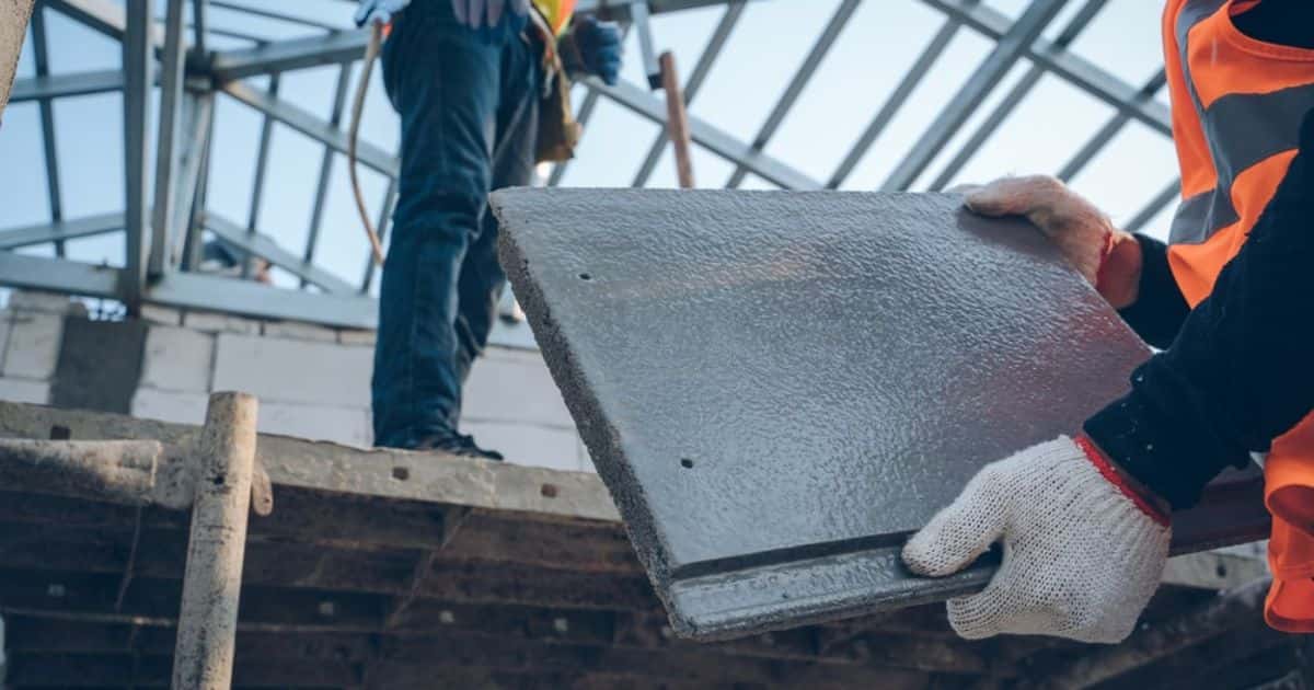Een dakwerker met fluohesje en werkhandschoenen houdt een betonnen dakpan vast: een soort dakbedekking met een levensduur van 30 tot 50 jaar.