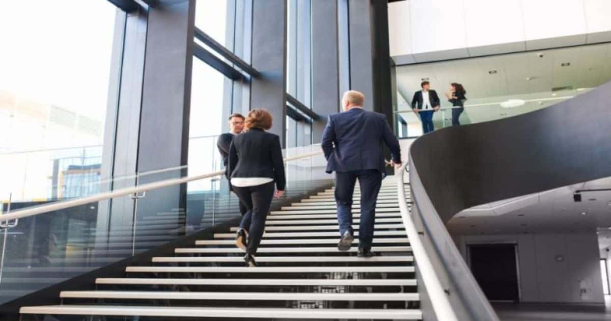 Grote open luie trap met een bocht waar 3 personen op stappen/staan. De trap bevindt zich in een publiek of kantoorgebouw.
