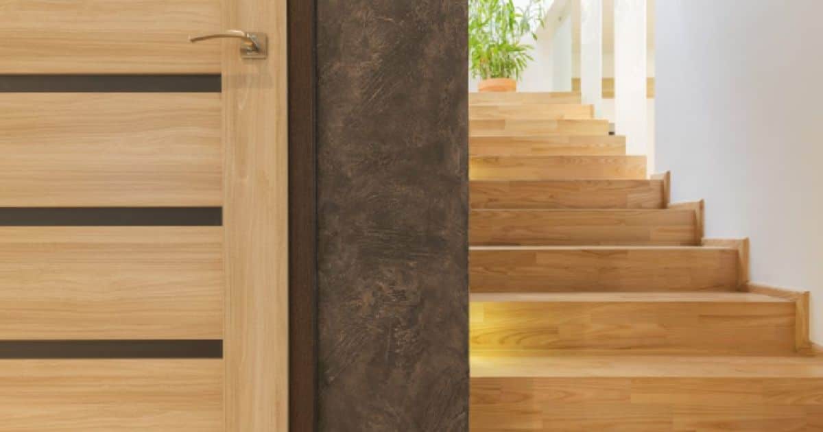 Rechte houten binnentrap, bovenaan de trap staat een plant. Er is ook een deur naast de trap. 