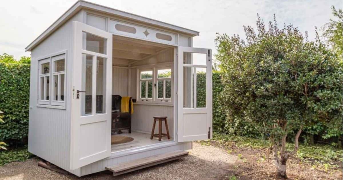 Een wit tuinhuis met een plat dak en dakbedekking. Voor struiken en naast een kleine boom.