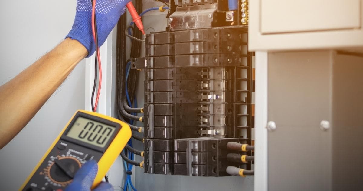 Een elektricien gebruikt een spanningsmeter bij de keuring van een meterkast. Hij draagt blauwe werkhandschoenen.