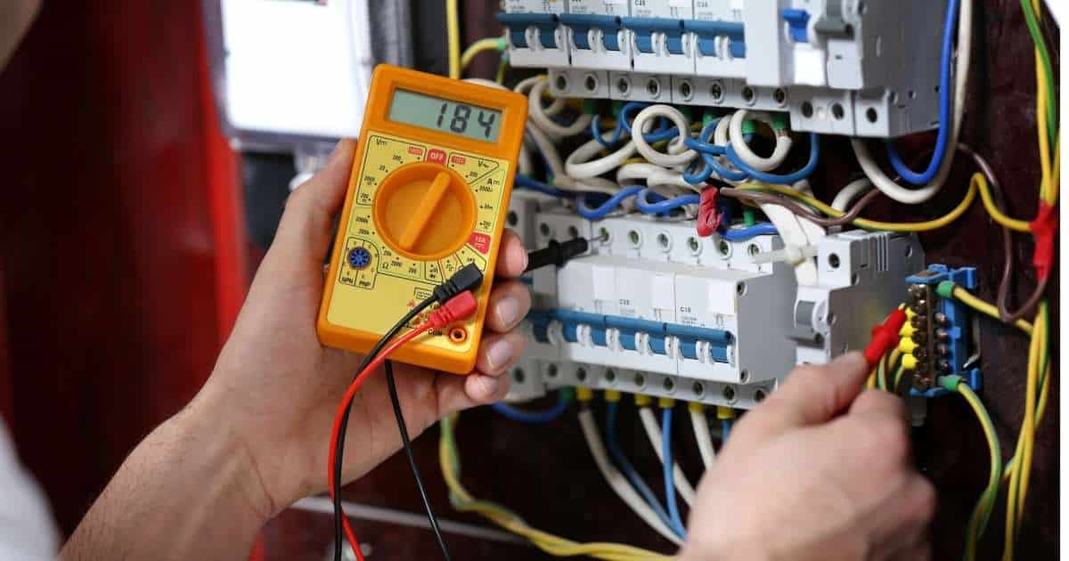 Twee handen in een meterkast. In de linkerhand een oranje spanningsmeter die het cijfer 184 weergeeft. In de rechterhand een plug die tegen een van de zekeringen wordt gehouden.