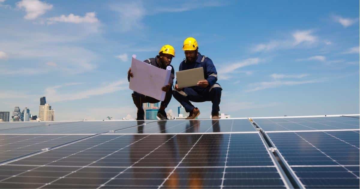 Twee elektriciens zitten gehurkt op een dak met zonnepanelen. Ze dragen gele veiligheidshelmen en werkkledij. De linkse keurder heeft een half opgerold plan vast. De rechtse heeft een tablet in de hand.