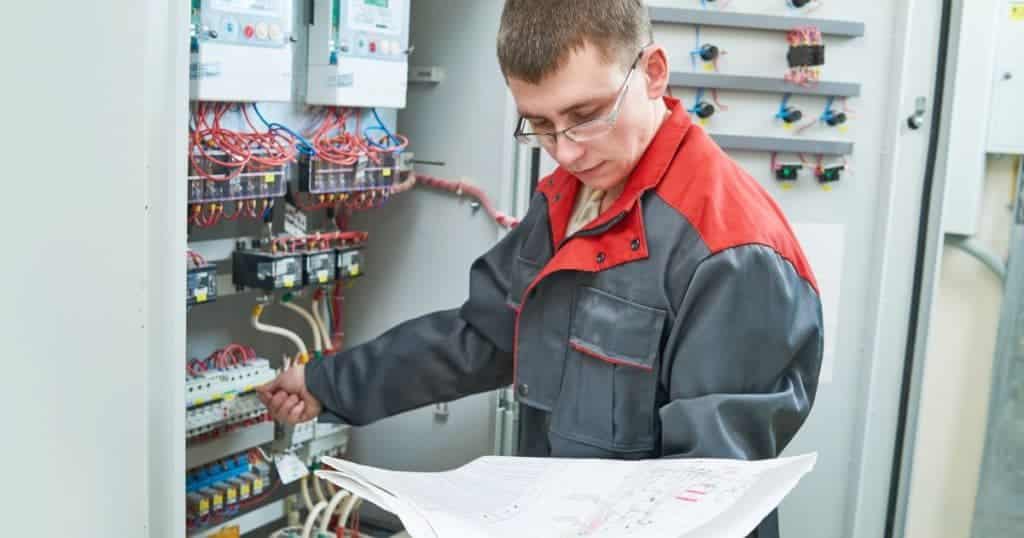 Een erkend keurder controleert een meterkast na de installatie van een thuisbatterij. Hij draagt een bril en een grijs-rode overal. Hij heeft een eendraadschema of situatieplan vast.