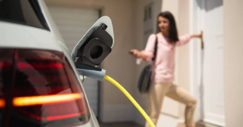 Een witte elektrische auto wordt opgeladen met een gele laadkabel . Op de achtergrond staat een vrouw met een roze blouse en beige broek. De vrouw gebruikt de sleutel om de auto te vergrendelen terwijl ze de deur van haar woning opent.