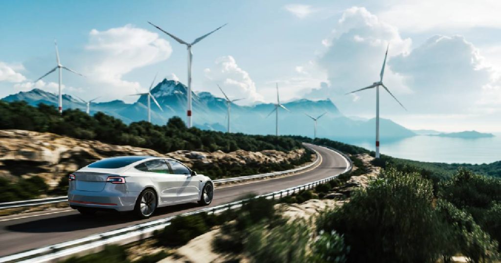 Een elektrische wagen rijdt op een weg die in een bocht loopt, op de achtergrond staan er windmolens, bergen, een meer en wolken.
