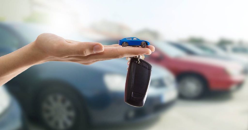 Een vrouw heeft een kleine elektrische wagen vast in haar hand, daaronder hangt een bos autosleutels. Op de achtergrond zien we enkele geparkeerde auto's
