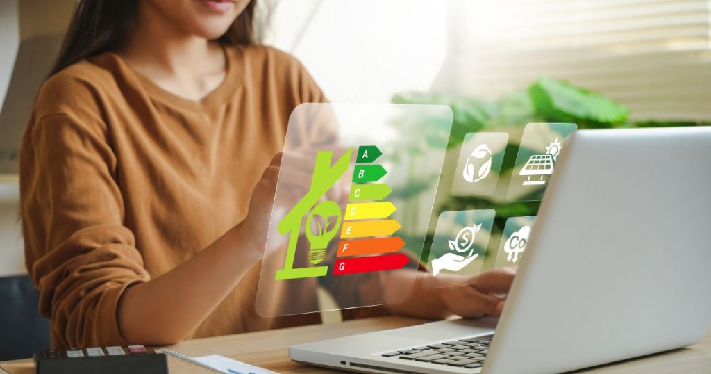 Een dame in bruin-oranje trui werkt op een lichtgrijze laptop. Ze bestudeert de energieaudit van haar woning? EPC, rendement zonnepanelen enzovoort.