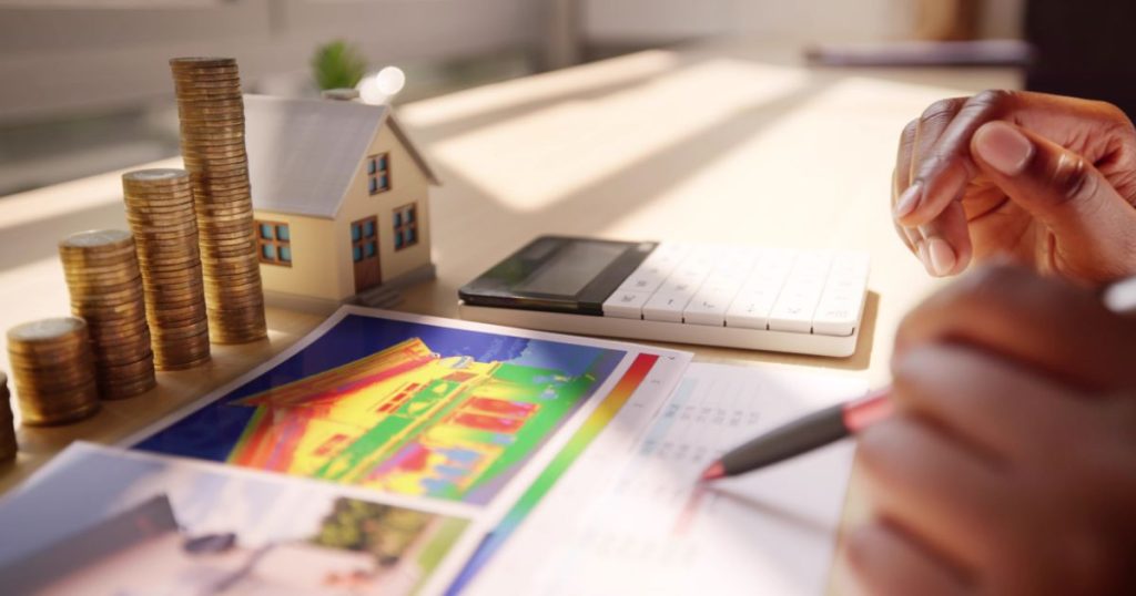Een hand richt een pen op een blad met allerhande cijfergegevens, daarboven een foto van de woning en een foto van de energiescan. Naast de papieren liggen een rekentoestel, een replica van het huis en munten.