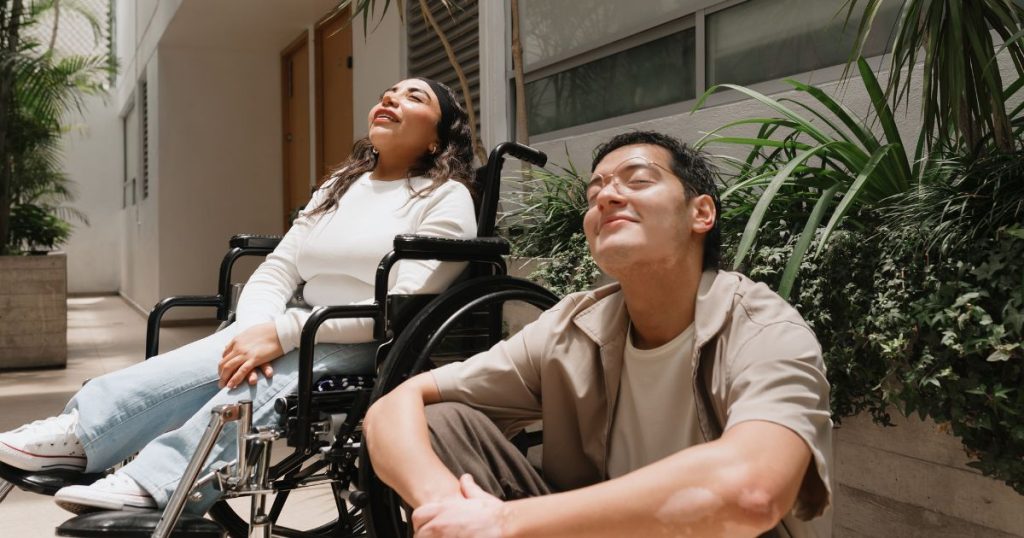 Een ergotherapeut in khaki hemd en met een ronde bril zit op de grond naast zijn patiënte die in een rolstoel zit en een wit t-shirt en een lichte jeansbroek draagt.