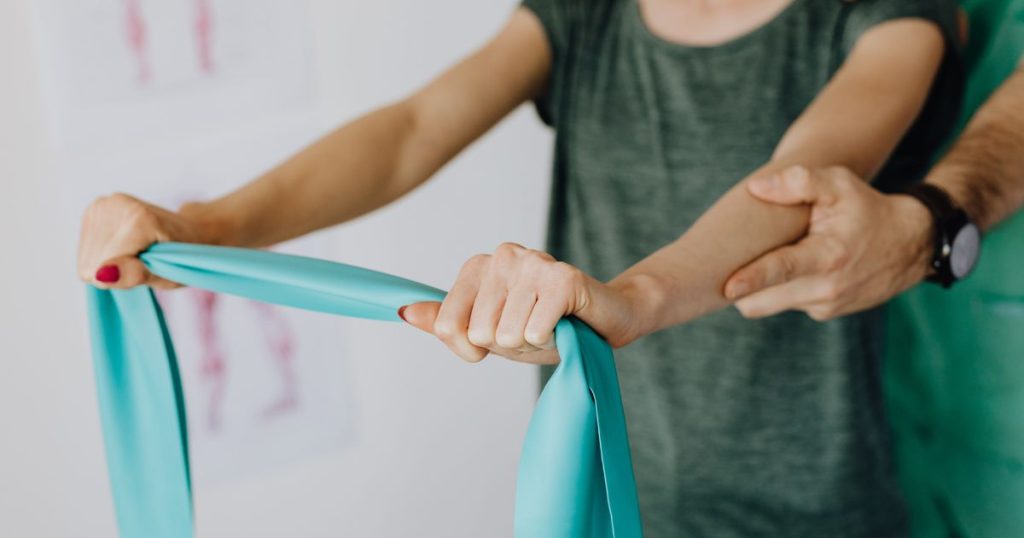 Een groenblauwe sjaal, strak gespannen tussen twee handen met rood gelakte nagels tijdens een sessie ergotherapie. Zichtbaar zijn de armen en het groene t-shirt van de patiënte en de arm met horloge en het groene tenue van de ergotherapeut.
