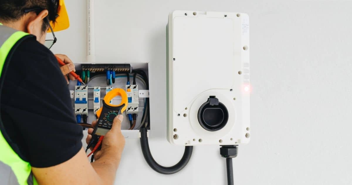 Een erkend keurder gebruikt een spanningsmeter tijdens de keuring van een witte, aan de muur gemonteerde laadpaal. Hij draagt een zwarte bril, gele veiligheidshelm, zwart t-shirt en fluohesje.