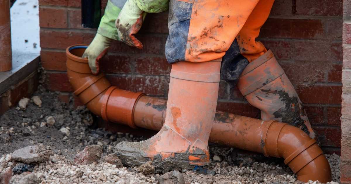 Een ontstoppingsspecialist heeft net een oranje ondergrondse leiding opgegraven om de oorzaak van een verstopping op te sporen. Hij draagt oranje laarzen, een oranje werkbroek, en groengele werkhandschoenen.