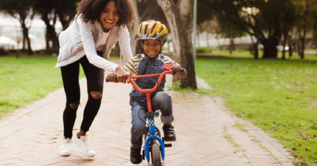 Een jonge moeder ondersteunt haar zoontje met gele helm bij het fietsen in een park, beschermd door hun familiale verzekering.