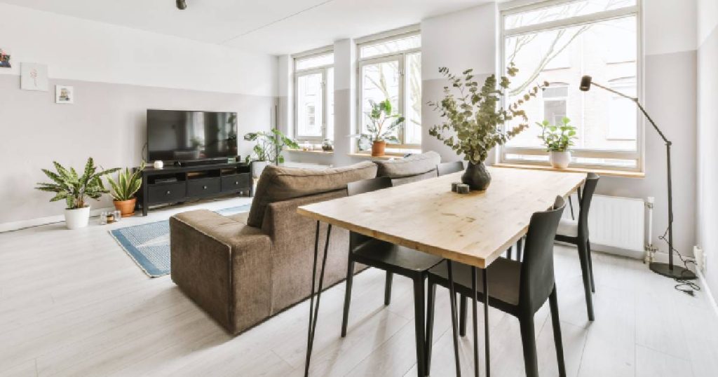 Een moderne living met eetkamer op lichtgrijs fineerparket. In de ruimte staan een houten tafel op ijzeren poten met zwarte stoelen, een lederen zetel, een lichtblauw tapijt, een televisie, verschillende kamerplanten en een zwarte lamp. De kamer heeft grote ramen waardoor veel licht binnenvalt.