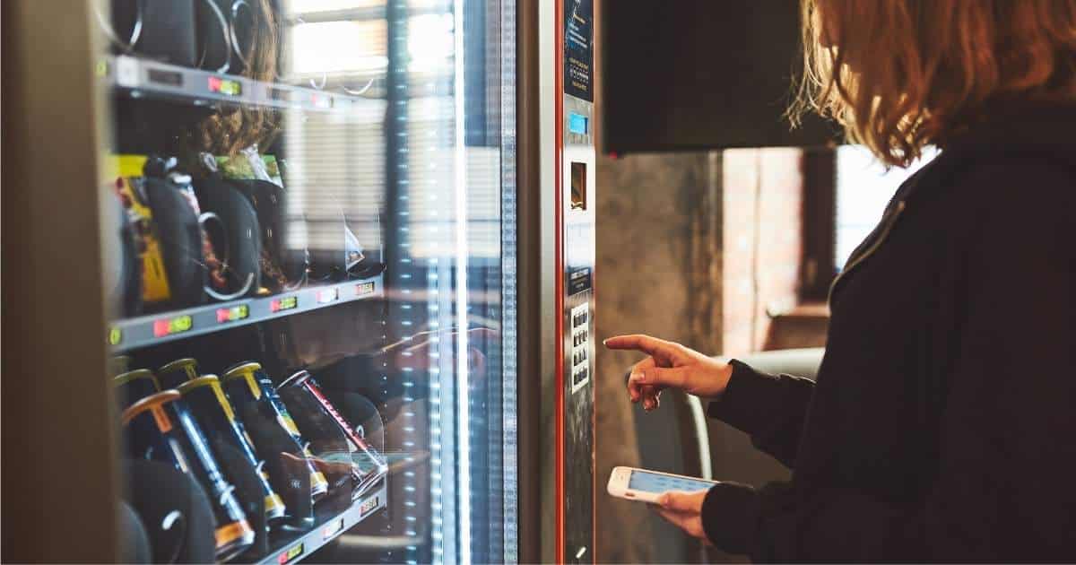 Een dame gebruikt het bedieningspaneel om een product te selecteren in een vendingmachine met dranken en snacks. Ze houdt haar smartphone vast om contactloos te betalen.