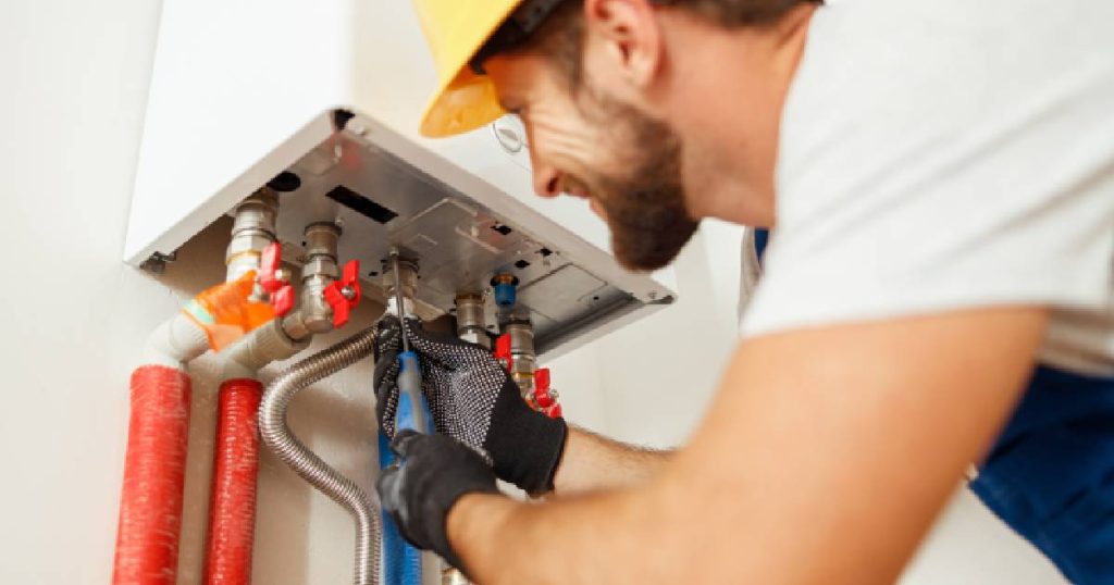 Een man in wit t-shirt, blauwe overall en gele veiligheidshelm controleert en onderhoudt een gascondensatieketel in een witte ruimte.