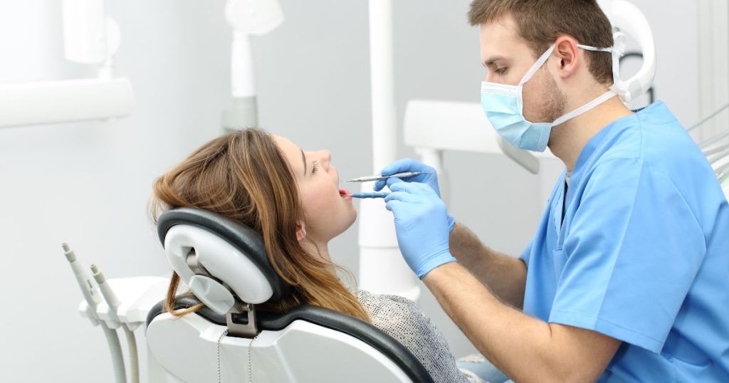 Een tandarts met blauwe tenue, blauw mondmasker en blauwe handschoenen behandelt zijn patiënte.