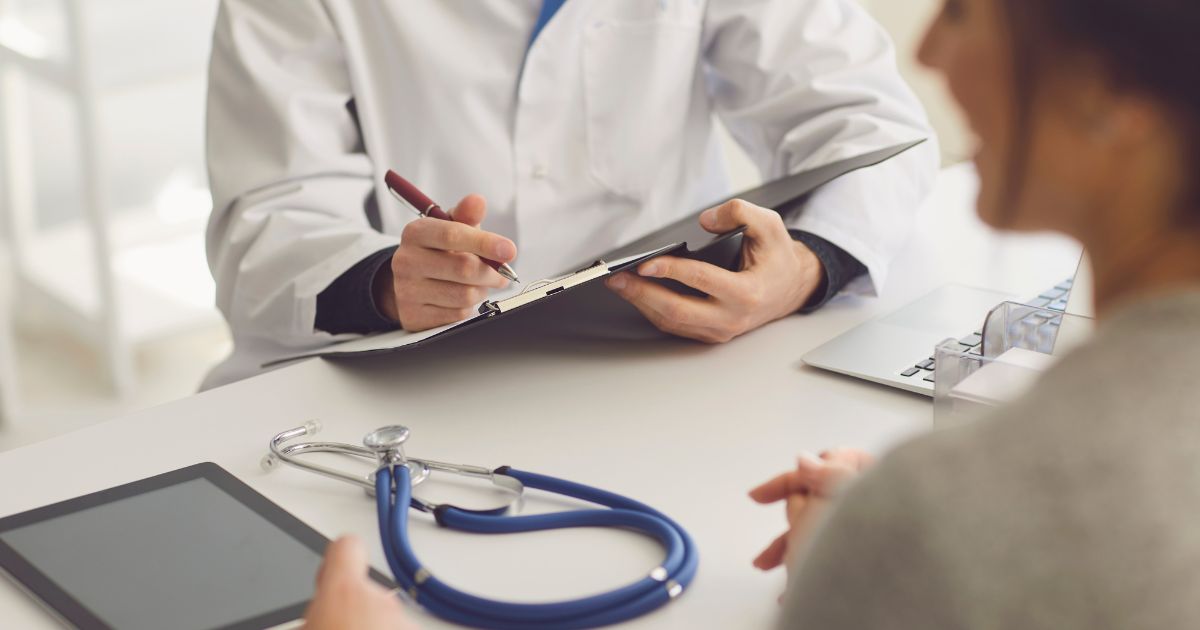 Een dokter bespreekt de gezondheidsstatus van de patiënt. De arts schrijft op een blad dat op een klembord ligt. Op de tafel liggen een tablet, een stethoscoop en een laptop.