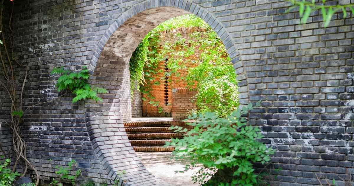 Een fraaie gemetselde tuinmuur met een grote, ronde opening uit grijze baksteen in Vlaams verband. De opening geeft uit op een terras met bakstenen trap, omringd door gemetselde muren.