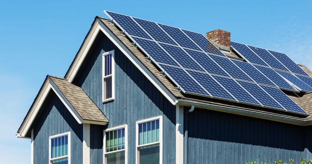Een woning met een blauwe houten gevel met verticale latten en witte raamkozijnen, een leien dak, een schoorsteen en een zonnepaneelinstallatie met 20 zonnepanelen.