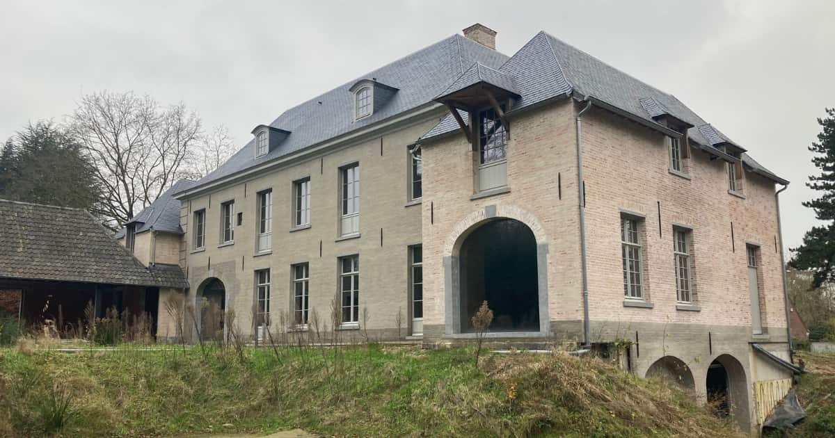 Een grote gerenoveerde Vlaamse hoeve met lichte bakstenen gevel, grijs leien dak met dakkapellen en een houten overkapping boven de rondboogingang staat op een bewolkte dag met verwilderde tuin op de voorgrond