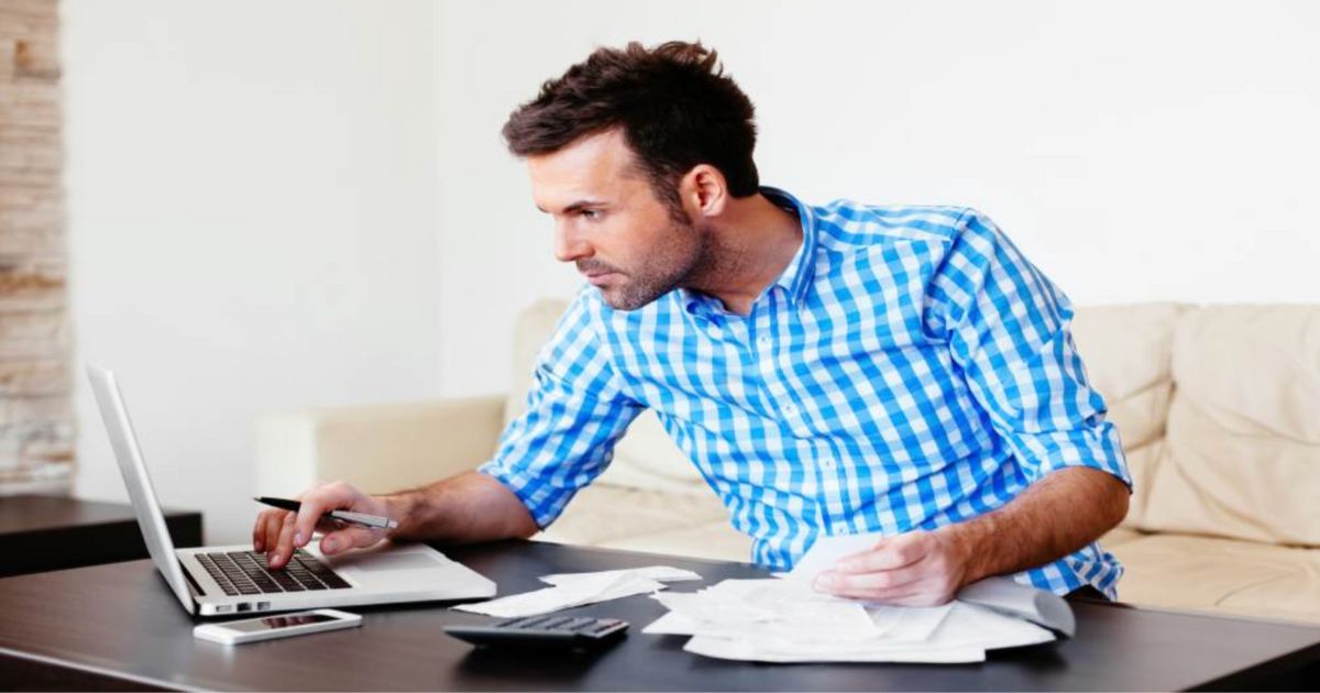 een man met een blauwe blouse zit aan een bureau te werken en heeft een laptop, telefoon, rekenmachine en stapel papieren voor zich