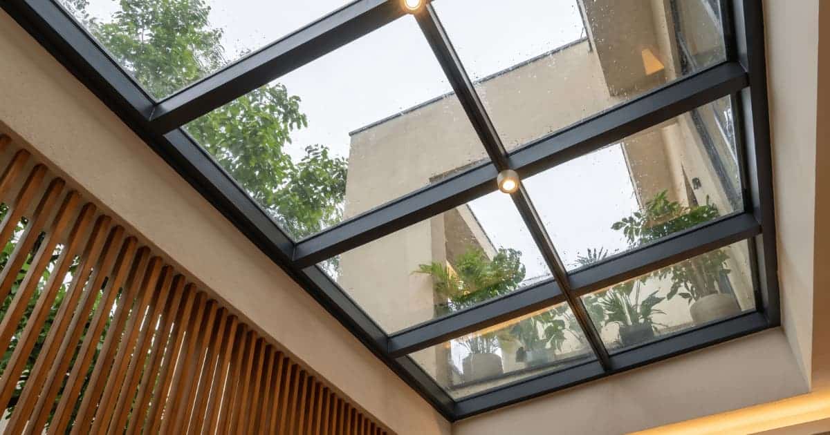 Onderaanzicht van een schuin glazen dak met transparante panelen in een donker antraciet metalen rasterframe waar regendruppels en groene beplanting doorheen zichtbaar zijn terwijl aan de linkerzijde een wand met verticale houten latten en warme inbouwspots het geheel afwerken.