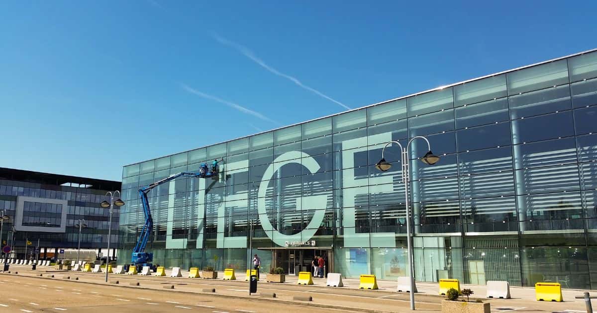 Een glazen bedrijfs- of kantoorgebouw op de luchthaven van Luik. Op de ramen staan de letters: Liège. Een man is met een blauwe kraanlift de ramen of glazen aan het poetsen.