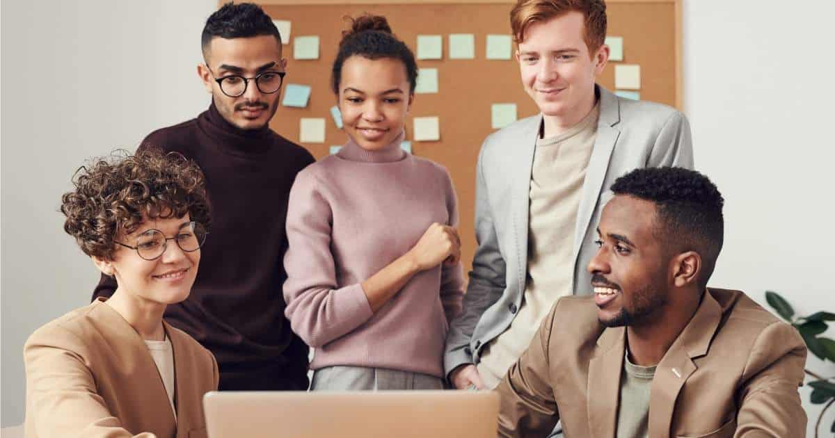 2 dames en 3 heren kijken naar een computerscherm. Ze glimlachen en de zittende man rechts kijkt naar zijn vrouwelijke collega.