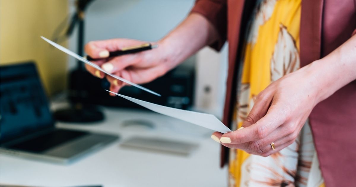 Ee dame met lichtgele nagellak in een rode blazer boven een jurk met geel en bruin bloemenmotief heeft in elke hand een document vast. In haar rechterhand heeft ze ook een zwarte pen. Op de achtergrond, vervaagd, een werktafel met laptop, bureaulamp en documenten.