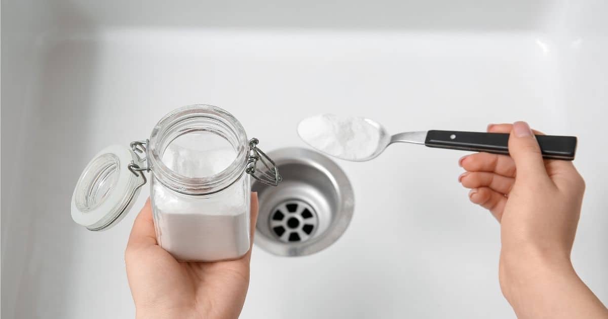 Twee handen houden een potje en een lepel zuiveringszout boven een witte gootsteen.