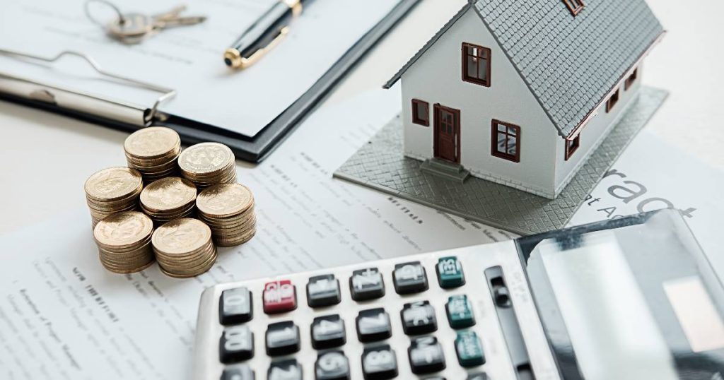 Symbolische voorstelling van de financiën van een huishouden: een rekenmachine, modelhuisje, stapel munten, pen en papieren.