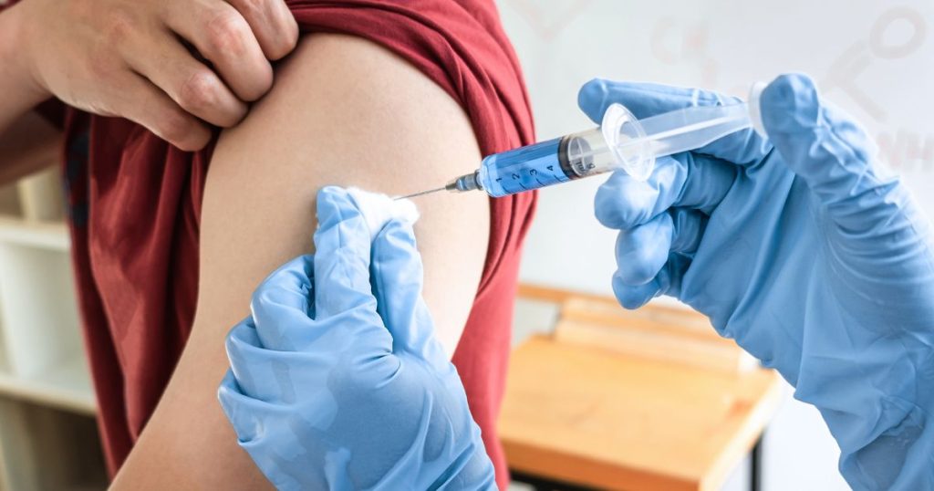 Een arts injecteert het griepvaccin in de arm van een jonge man met rood t-shirt. De arts draagt blauwe latex handschoenen en drukt een watje tegen de injectieplek.
