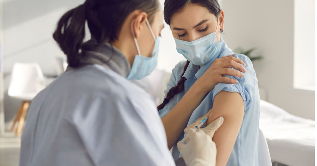 Een arts injecteert het griepvaccin in de arm van een jonge vrouw. Ze dragen beiden een blauw hemd en blauw mondmasker.