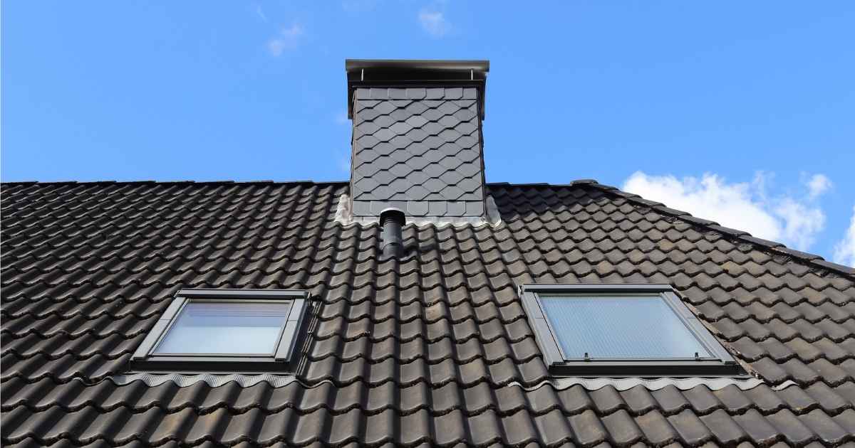Ee dak met bruine dakpannen, een schouw met platte dakpannen, en 2 kleine dakramen tegen een helderblauwe hemel.