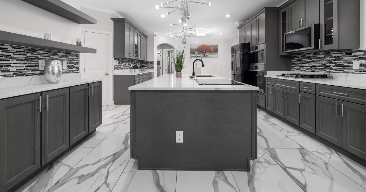 Een moderne grijs met witte keuken met keukeneiland. De keuken heeft een tegelvloer met marmeren natuurstenen tegels. Op de grijze houten aanrechtblokken liggen witte natuurstenen aanrechtbladen.