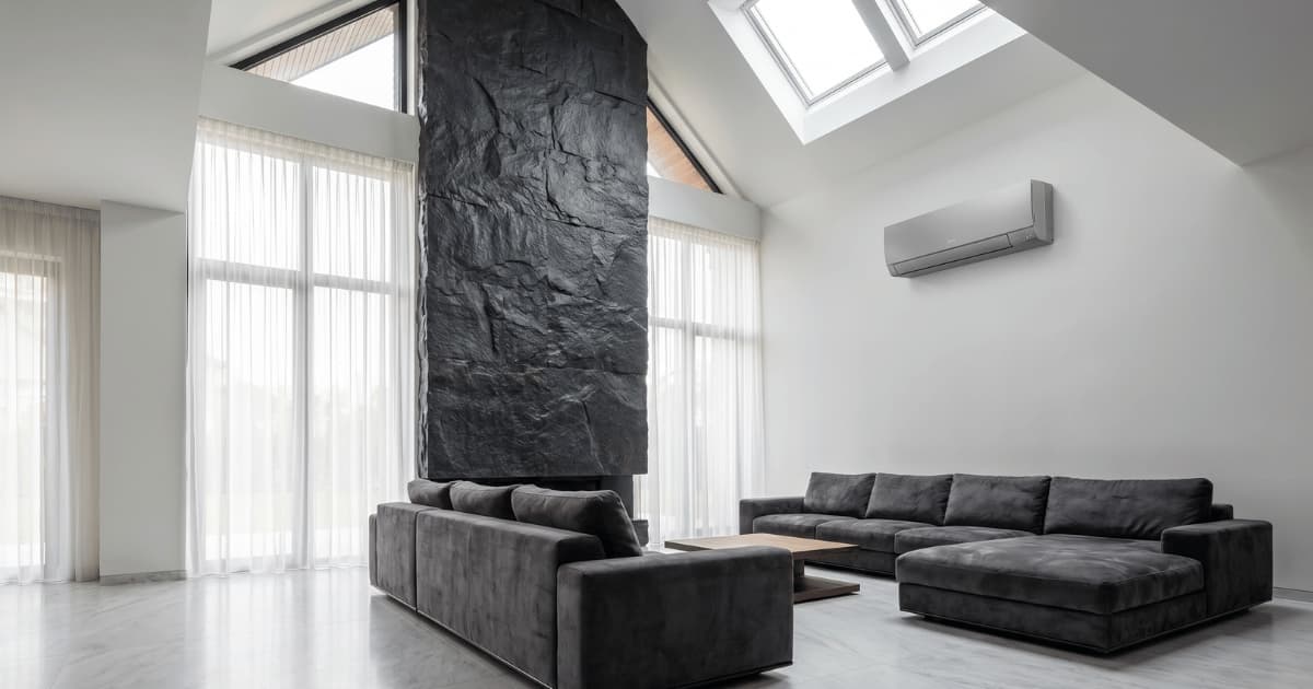Een grijze airco-binnenunit hangt hoog aan een witte muur in een ruime woonkamer met dubbelhoog plafond, dakramen, een donkere natuurstenen zuil en twee antracietkleurige hoekbanken op een lichtgrijze vloer.