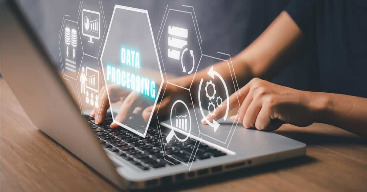 Een grijze laptop op een houten tafel, met de handen van een vrouw in zwart t-shirt op het toetsenbord. Voor het scherm en boven het toetsenbord zweven verschillende hologramachtige zeshoeken. De grootste bevat de woorden 'data processing', de andere zeshoeken bevatten symbolen en iconen.