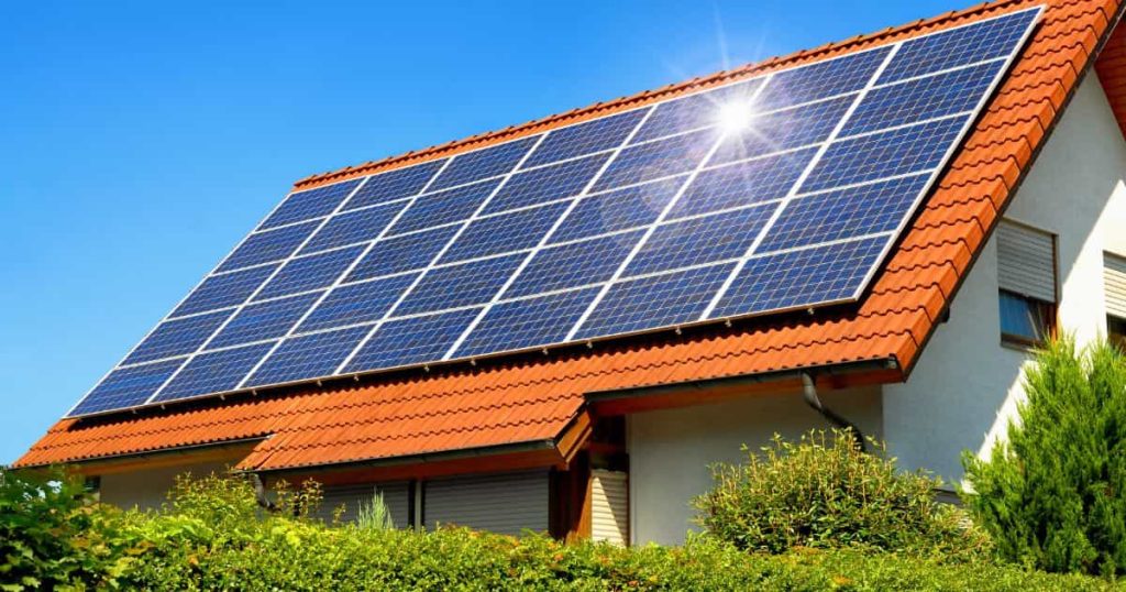 Een huis met een oranje dak en witte muren. Het dak is bedekt met zonnepanelen waar de zon fel op schijnt.