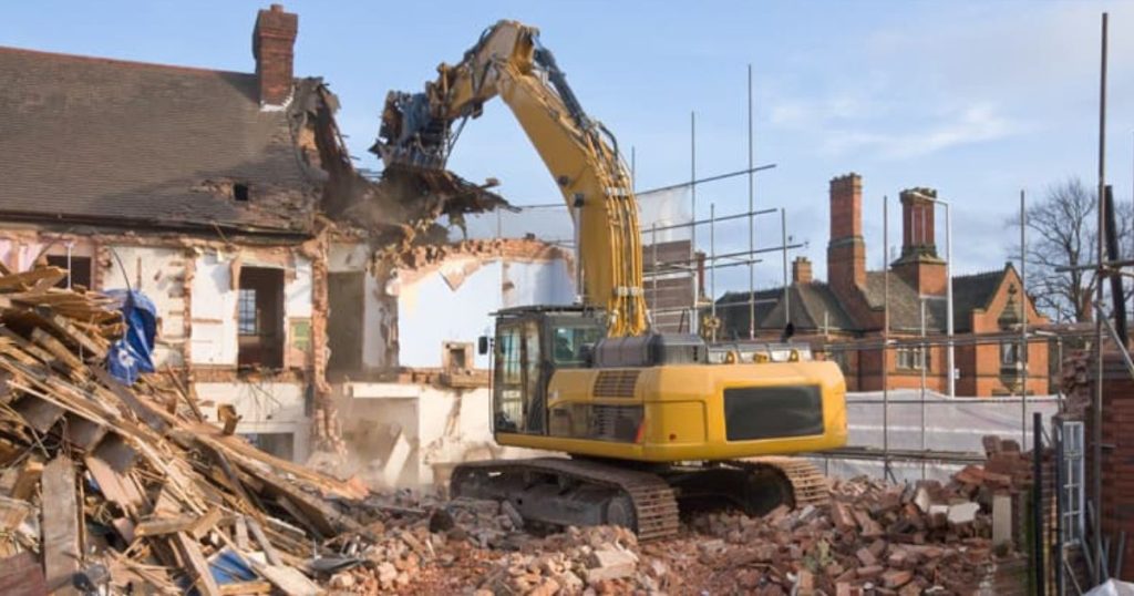 Een graafmachine sloopt een ouder gebouw met leien dak. Rondom de woonst staat een stellingenconstructie. De woonst wordt met de grond gelijk gemaakt.