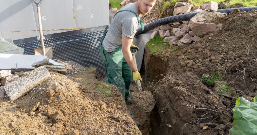 Een vakman in groene werkoverall is met een metalen schop bezig met het graven van een geul: voor afvoerleidingen of voor bekabeling?