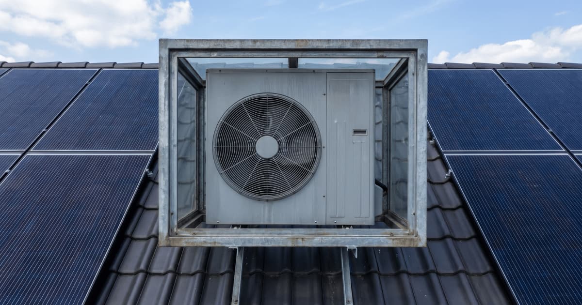 Grote grijze airco-buitenunit in een metalen frame op de nok van een donker pannendak met aan weerszijden rijen donkerblauwe zonnepanelen tegen een blauwe lucht.