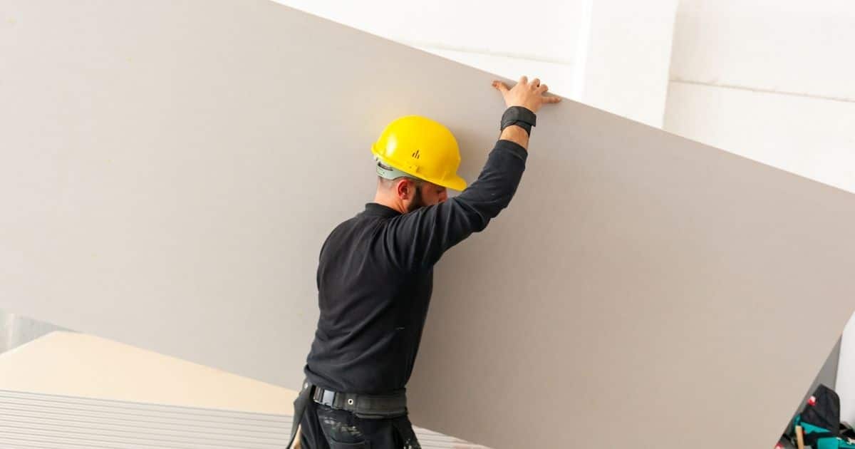 Een gyprocspecialist draagt een grote, gebroken witte gyprocplaat. De specialist draagt een zwart t-shirt, zwarte broek, gereedschapsriem en gele veiligheidshelm.