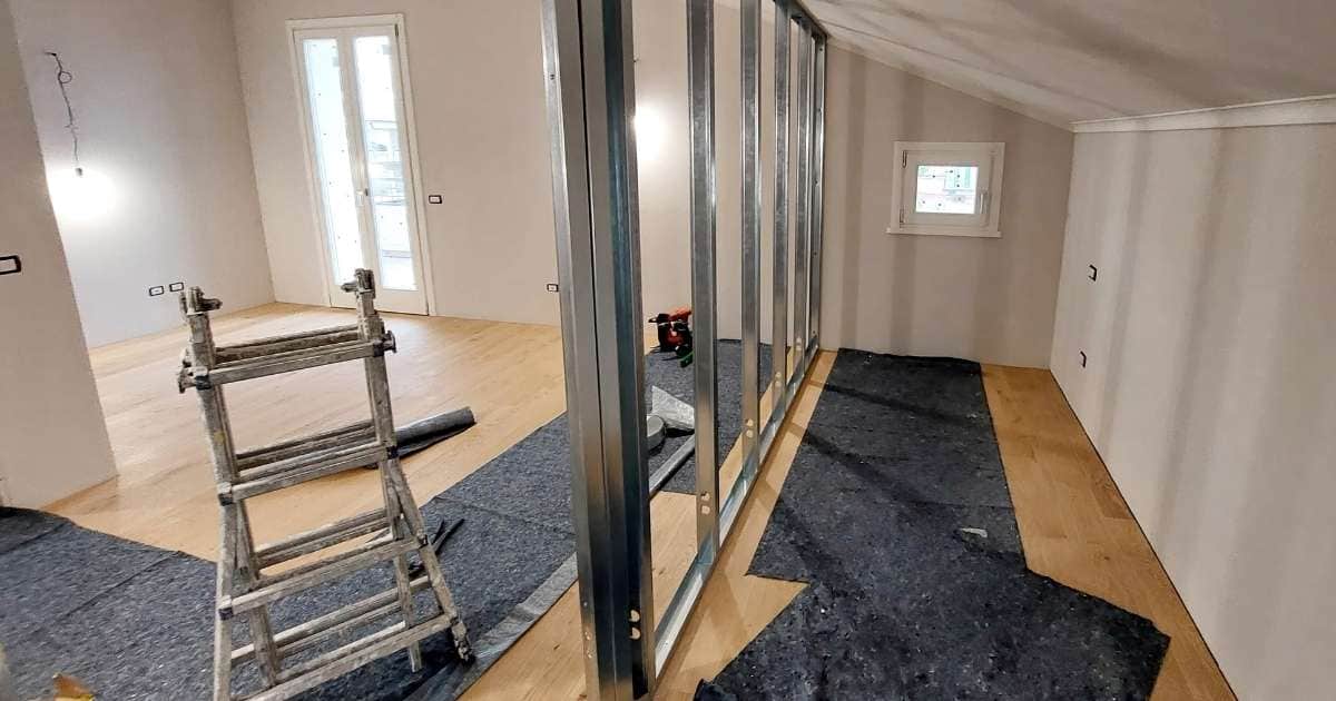 Een gerenoveerde ruimte die wordt afgewerkt met gyprocplaten. De ruimte wordt verdeeld met een scheidingswand: er staat al een metalen frame klaar waar de gyprocplaten in bevestigd zullen worden. De ruimte heeft een houten vloer waarop beschermmatten werden gelegd.