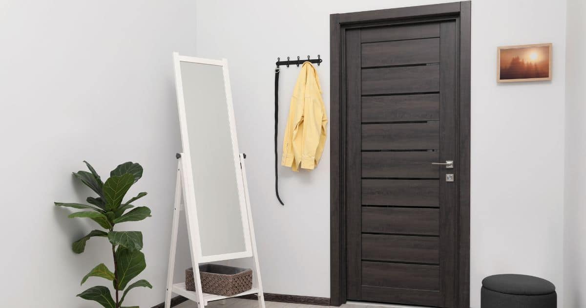 Hal in gebroken wit met donkere houten deur. In de ruimte staan een witte spiegel, een plant en een poef. Rechts van de deur hangt een kapstok aan de muur.