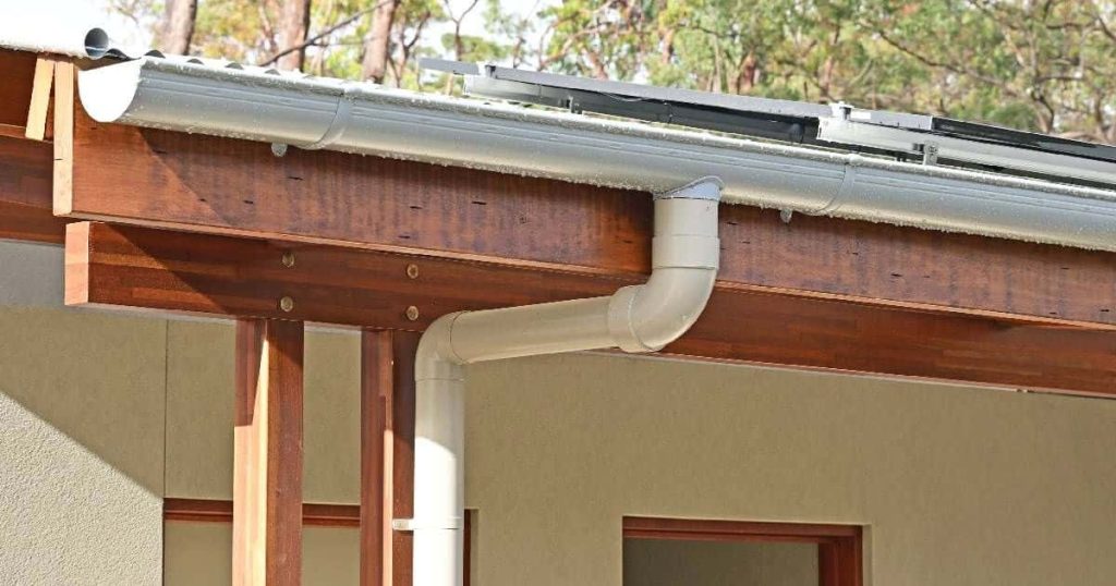 Een halfronde dakgoot of hanggoot bevestigd aan een houten balk en het uiteinde van een dak met zonnepanelen.